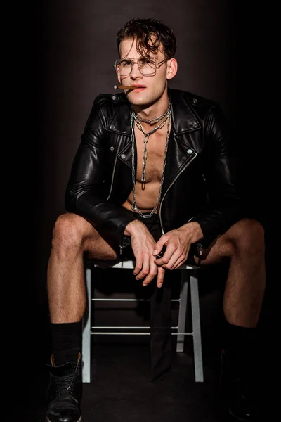 Brutale uomo in giacca di pelle seduto sulla sedia con sigaro su nero — Foto stock