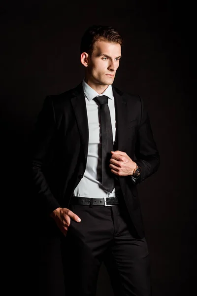 Hombre de negocios guapo en ropa formal de pie con la mano en el bolsillo aislado en negro - foto de stock