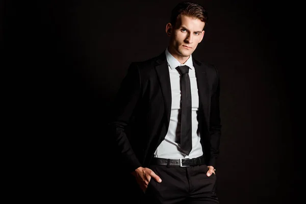 Hombre de negocios guapo en ropa formal de pie con las manos en bolsillos aislados en negro — Stock Photo