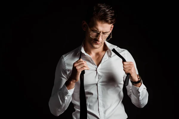 Guapo hombre de negocios en gafas quitándose la corbata aislado en negro - foto de stock