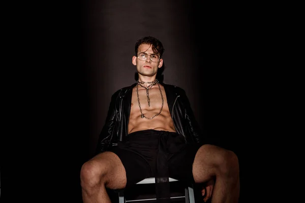 Сексуальный и жестокий молодой человек в очках и кожаной куртке, сидящий на черном — стоковое фото