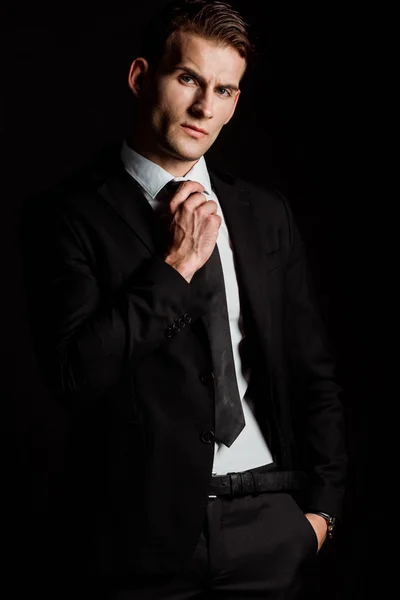 Handsome businessman man touching tie while standing with hand in pocket isolated on black — Stock Photo