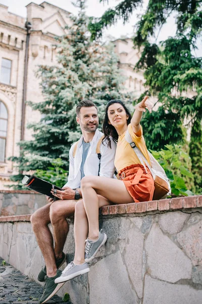 Mujer atractiva señalando con el dedo y sentado cerca de hombre feliz con mapa - foto de stock