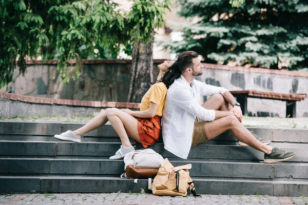 Seitenansicht eines bärtigen Mannes und einer fröhlichen Frau, die auf Treppen in der Nähe von Rucksäcken sitzen — Stockfoto