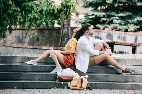 Vista lateral del hombre barbudo y la mujer alegre sentado en las escaleras cerca de mochilas - foto de stock