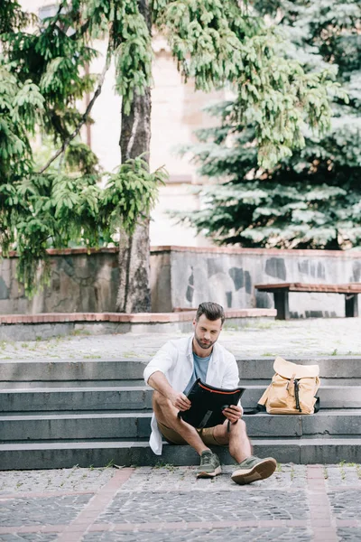 Handsome bearded man sitting on stairs and looking at map near trees — Stock Photo