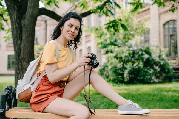 Menina alegre segurando câmera digital enquanto sentado no banco perto da universidade — Fotografia de Stock