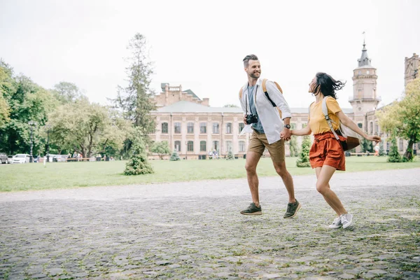 Cheerful bearded man and woman holding hands while running near building — Stock Photo