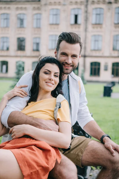 Alegre pareja mirando la cámara mientras está sentado cerca de la universidad - foto de stock