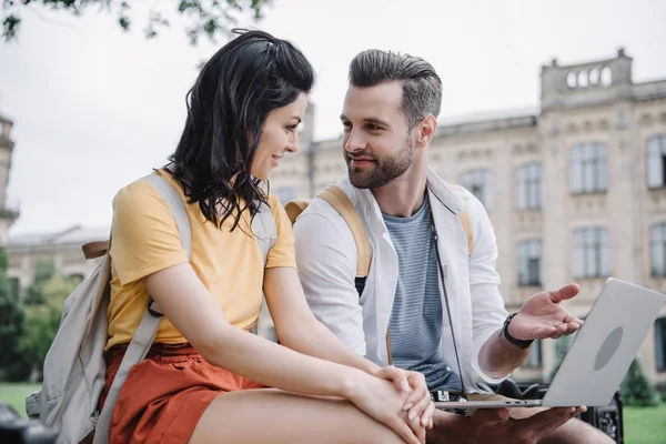 Feliz joven sentada cerca del hombre barbudo usando el ordenador portátil - foto de stock