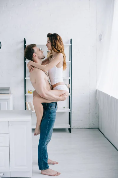 Vista lateral de novio guapo sosteniendo novia atractiva en apartamento - foto de stock