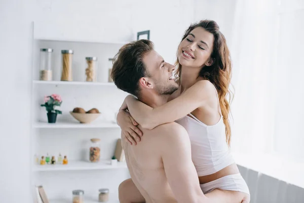 Guapo novio sosteniendo sonriente y atractiva novia en apartamento - foto de stock