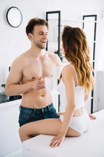 Novia y novio guapo sonriendo y sosteniendo tazas con té en el apartamento - foto de stock