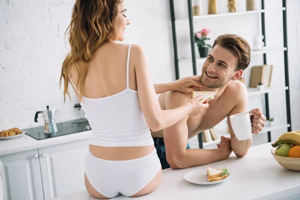 Back view of girlfriend feeding with sandwich handsome boyfriend in apartment — Stock Photo