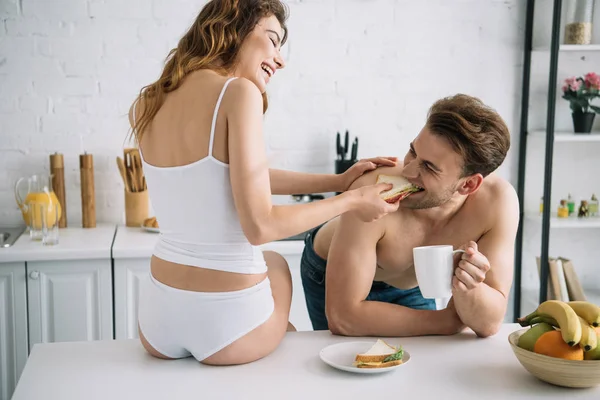 Back view of girlfriend feeding with sandwich handsome boyfriend in apartment — Stock Photo