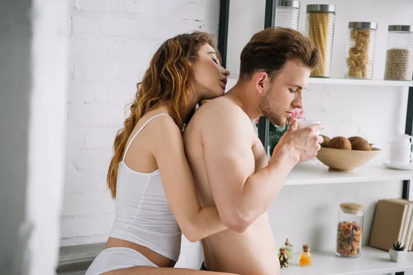 Attractive girlfriend hugging handsome boyfriend with cup in apartment — Stock Photo