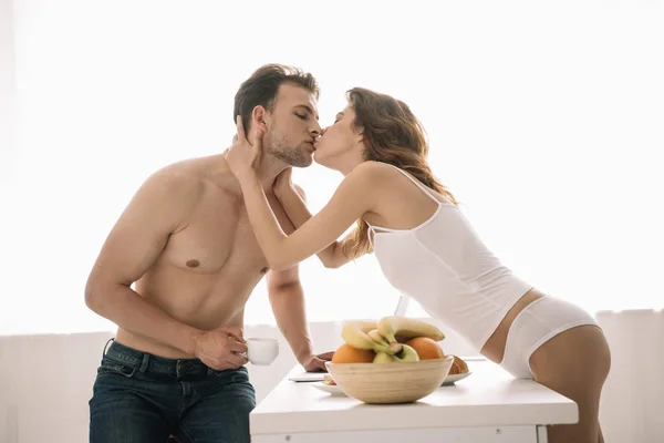 Attractive woman kissing and hugging handsome man with cup in apartment — Stock Photo