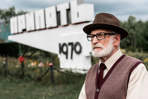 ПРИПЯТ, УКРАИНА - 15 августа 2019 года: старший бородатый мужчина в очках стоит у памятника с припятскими буквами — Stock Photo