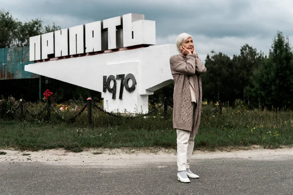Прип'ять, Україна-15 серпня 2019: відставна жінка стояла біля пам'ятника з Прип'яті та накритими вухами — стокове фото