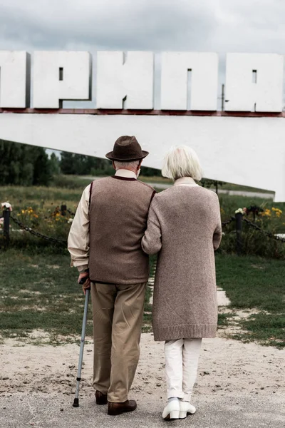 PRIPYAT, UCRÂNIA - AGOSTO 15, 2019: vista traseira do marido e da esposa aposentados que estão perto do monumento com letras — Fotografia de Stock