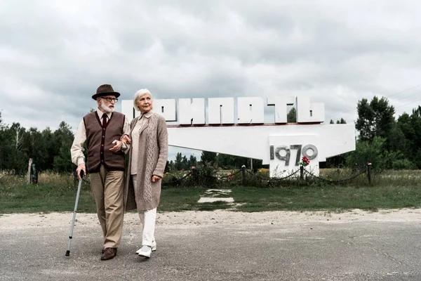 PRIPYAT, UKRAINE - 15 AOÛT 2019 : couple de personnes âgées marchant près d'un monument avec des lettres pripyat — Photo de stock
