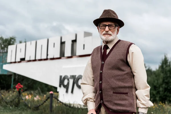 PRIPYAT, UCRÂNIA - AGOSTO 15, 2019: homem sênior de óculos em pé perto do monumento com letras pripyat — Fotografia de Stock