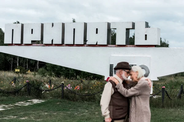 PRIPYAT, UCRAINA - 15 AGOSTO 2019: focus selettivo del monumento con lettere di pripyat vicino alla coppia anziana che si abbraccia all'esterno — Foto stock