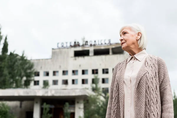 PRIPYAT, UKRAINE - AUGUST 15, 2019: low angle view of senior woman standing near building with hotel polissya lettering in chernobyl — Stock Photo