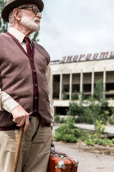 PRIPYAT, UKRAINE - AUGUST 15, 2019: senior man holding suitcase near building with energetic lettering in chernobyl — Stock Photo