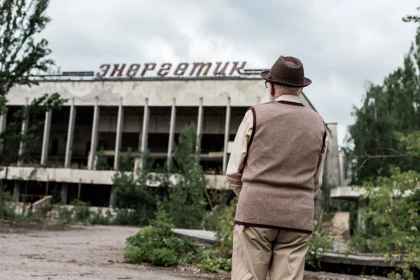 PRIPYAT, UKRAINE - 15 AOÛT 2019 : vue de dos d'un retraité coiffé d'un chapeau debout près d'un immeuble avec un lettrage énergique en tchernobyl — Photo de stock