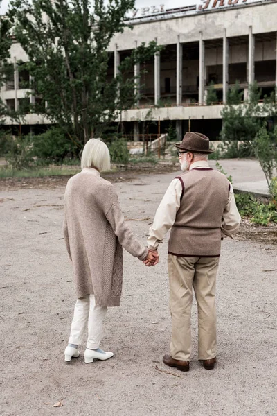 PRIPYAT, UCRANIA - 15 de agosto de 2019: vista trasera del hombre y la mujer jubilados tomados de la mano cerca del edificio - foto de stock