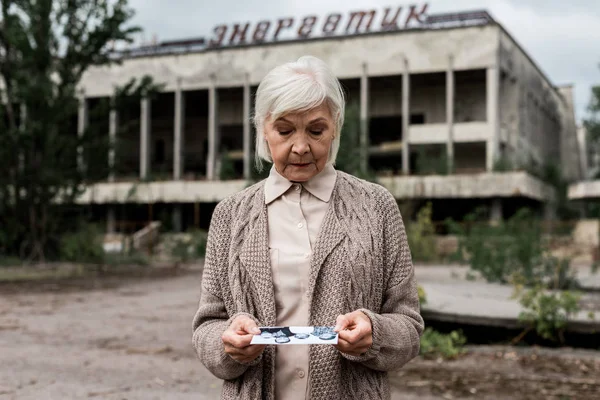 PRIPYAT, UKRAINE - AUGUST 15, 2019: senior woman looking at photo near building with energetic lettering in chernobyl — Stock Photo