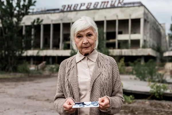 Pripjat, Ukraine - 15. August 2019: Seniorin mit Foto in der Nähe eines Gebäudes mit Schriftzug in Tschernobyl — Stockfoto
