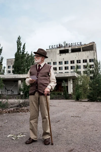 PRIPYAT, UCRAINA - 15 AGOSTO 2019: senior man with walking cane holding photo near building with hotel polissya letters — Foto stock