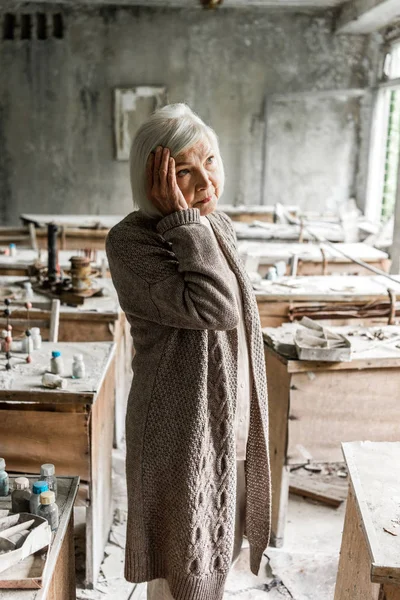 Mujer jubilada tocando el pelo gris y de pie en el aula sucia - foto de stock