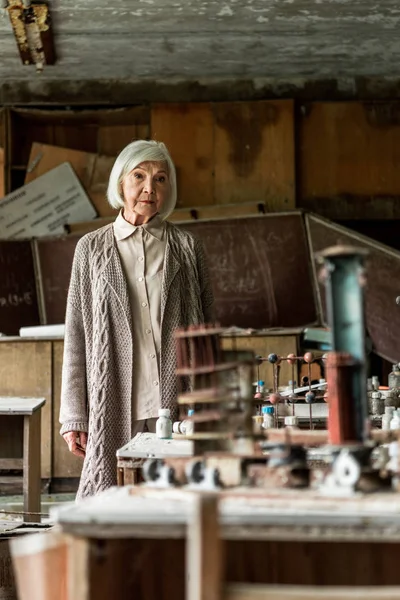 Foco seletivo da mulher aposentada em pé em sala de aula abandonada suja — Fotografia de Stock