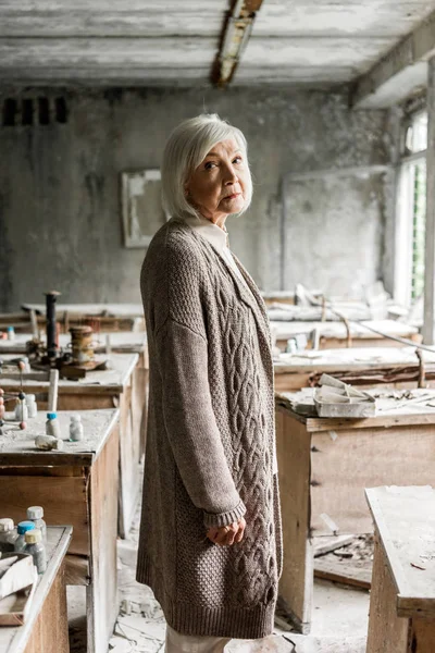 Selective focus of frustrated retired woman standing in dirty abandoned classroom — Stock Photo