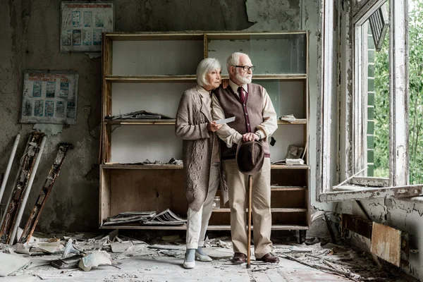 Homme et femme à la retraite regardant la fenêtre tout en se tenant dans la salle de classe endommagée — Photo de stock