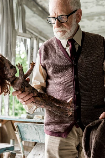 Bearded senior man holding hat while looking at burnt baby doll in classroom — Stock Photo