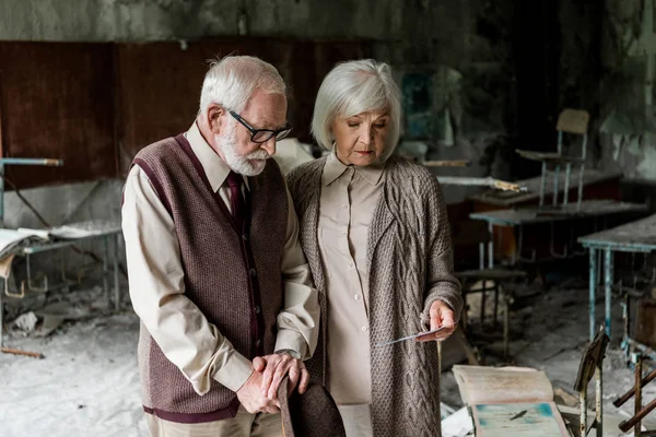 Retired couple looking at photo while standing in damaged classroom — Stock Photo