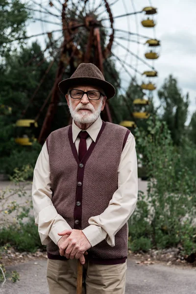 PRIPYAT, UKRAINE - AUGUST 15, 2019: bearded retired man in hat standing with walking cane in amusement park with ferris wheel — Stock Photo