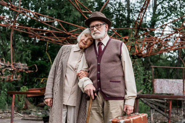 ПРИПЯТ, УКРАИНА - 15 августа 2019 года: старший мужчина в шляпе, держащий чемодан, стоя рядом с женой возле заброшенной карусели — стоковое фото