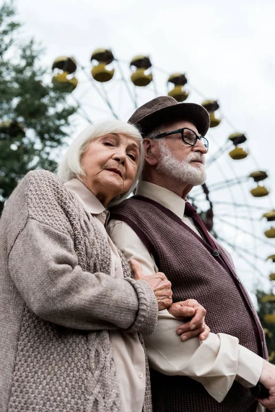 PRIPYAT, UCRAINA - 15 AGOSTO 2019: vista a basso angolo dell'anziano barbuto in occhiali con moglie vicino alla ruota panoramica nel parco divertimenti — Foto stock