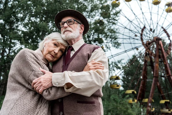 PRIPYAT, UCRANIA - 15 de agosto de 2019: un hombre mayor barbudo abrazándose con su esposa cerca de la rueda del hurón en el parque de atracciones - foto de stock