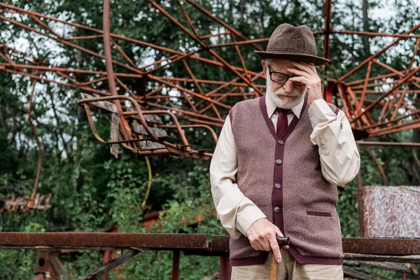 PRIPYAT, UKRAINE - 15 AOÛT 2019 : homme à la retraite en chapeau et lunettes debout avec canne à pied près du carrousel endommagé — Photo de stock