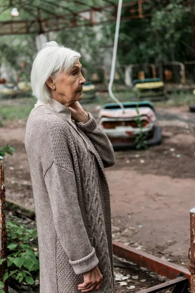 PRIPYAT, UKRAINE - 15 AOÛT 2019 : Femme âgée debout dans un parc d'attractions près de voitures tampons abandonnées — Photo de stock