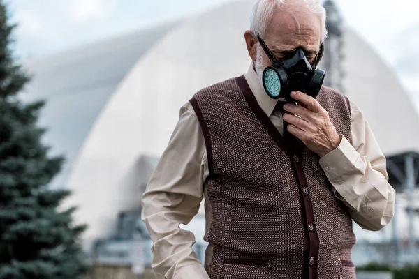 PRIPYAT, UCRÂNIA - AGOSTO 15, 2019: homem sênior tocando máscara protetora e de pé perto de reator chernobyl abandonado — Fotografia de Stock