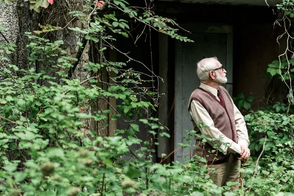 Selective focus of retired man in glasses standing with walking stick near house — Stock Photo