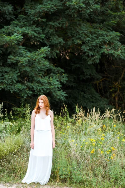 Красивая рыжая девушка в белом платье, стоящая в поле с полевыми цветами — стоковое фото