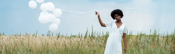 Tiro panorâmico de menina americana africana atraente no vestido segurando balões no campo — Fotografia de Stock